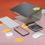 Apple Magic Keyboard, Magic Mouse, iPhone, AirPods case, calculator, and laptop on pink and red desk with yellow arrow sticky notes