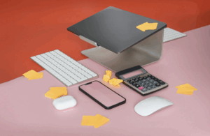 Apple Magic Keyboard, Magic Mouse, iPhone, AirPods case, calculator, and laptop on pink and red desk with yellow arrow sticky notes