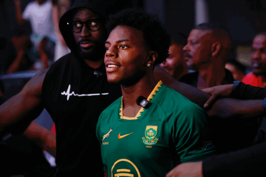 Young man wearing a South Africa rugby jersey with a microphone clipped on, surrounded by people in a dimly lit setting.