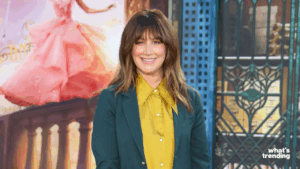 Woman with brown hair wearing a green blazer and yellow shirt smiling in front of a Cinderella poster