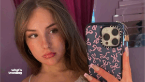 Young woman with long brown hair taking a mirror selfie with a phone case decorated with pink bows