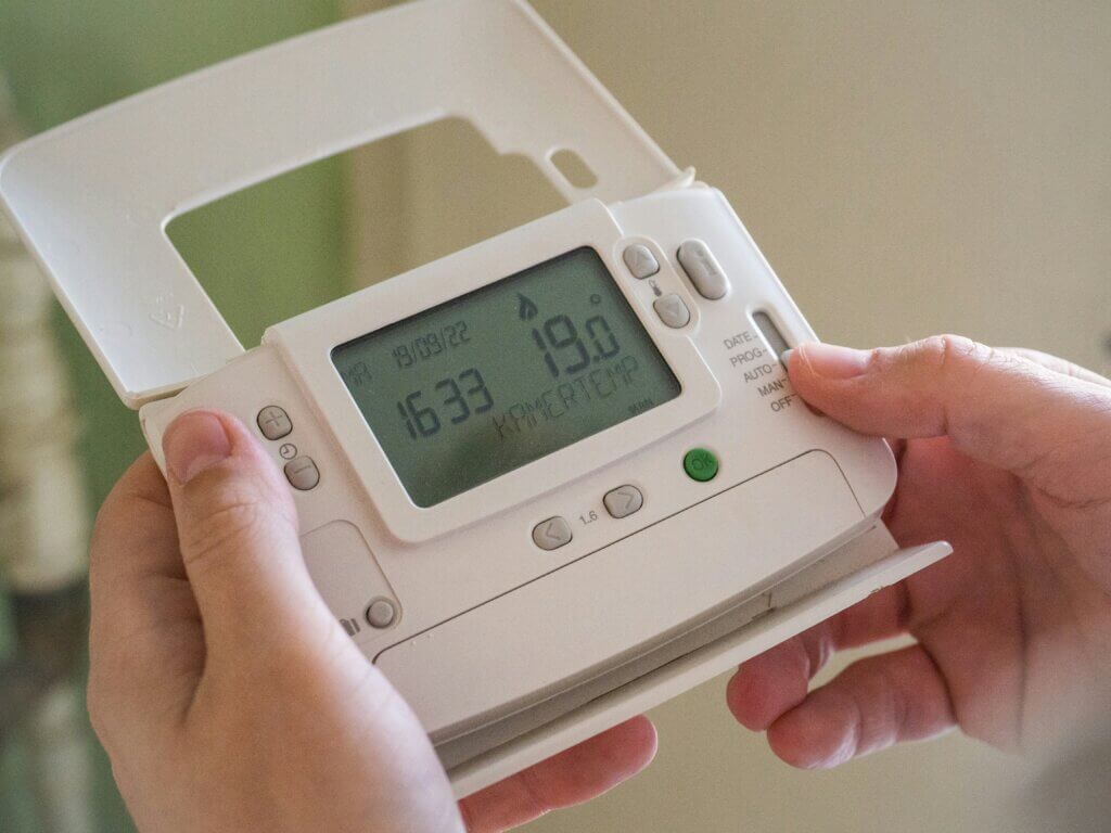 Hands adjusting a white digital thermostat displaying 19.0 degrees and the time 16:33 on 18/09/22