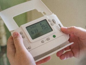 Hands adjusting a white digital thermostat displaying 19.0 degrees and the time 16:33 on 18/09/22