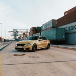 Gold BMW M4 parked on a concrete road near stacked shipping containers at a port terminal.