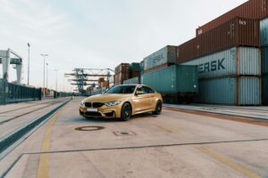 Gold BMW M4 parked on a concrete road near stacked shipping containers at a port terminal.