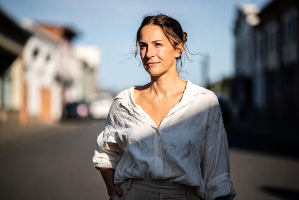 Woman with light brown hair in a white patterned blouse standing outdoors in sunlight on a blurred street background