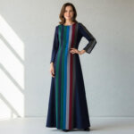 Woman wearing a long navy dress with vertical multicolored stripes standing against a light gray wall