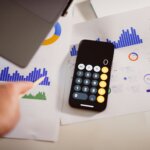 Hand pointing with a pencil at blue bar charts on paper next to a smartphone calculator app and laptop corner