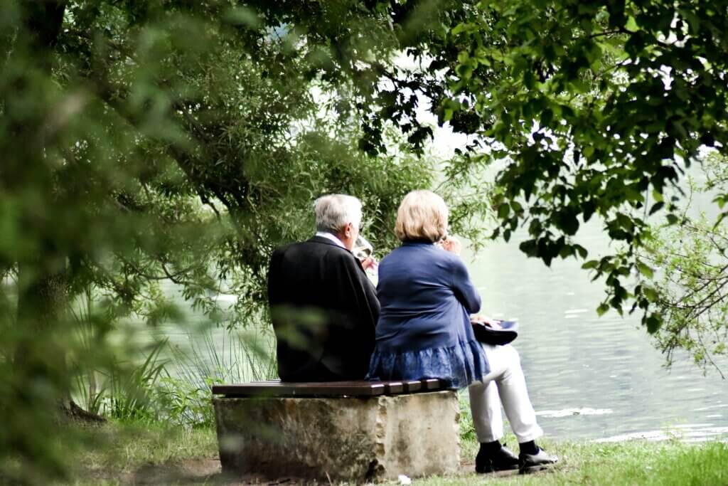 Elderly couple sitting on a bench by a lake, drinking from wine glasses surrounded by green trees.