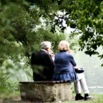 Elderly couple sitting on a bench by a lake, drinking from wine glasses surrounded by green trees.