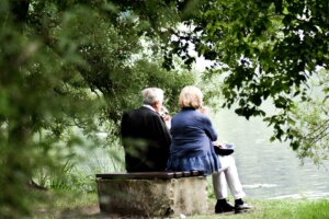 Elderly couple sitting on a bench by a lake, drinking from wine glasses surrounded by green trees.