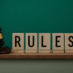 Wooden blocks spelling "RULES" next to a judge's gavel and two small wooden house models on a shelf