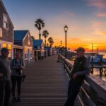 Three people talking on a wooden pier near fishing boats at sunset by buildings with a "Percy Stainly" sign