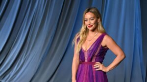 Hilary Duff wearing a purple pleated gown posing on a gray carpet with a blue draped backdrop