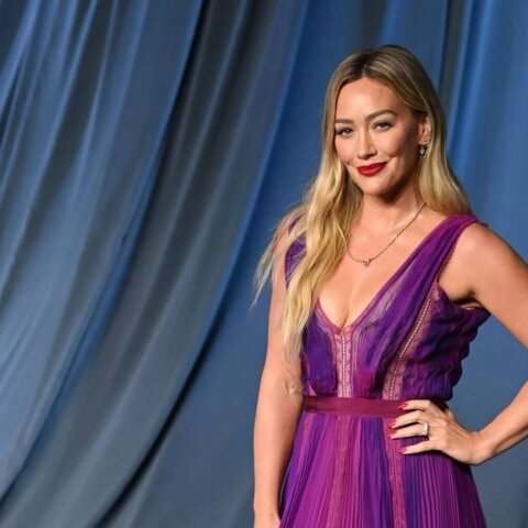 Hilary Duff wearing a purple pleated gown posing on a gray carpet with a blue draped backdrop