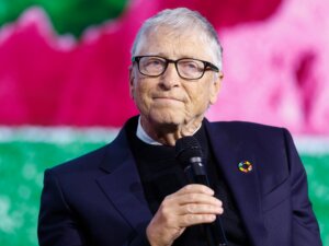 Bill Gates speaking into a microphone wearing glasses and a dark suit with a colorful pin on the lapel.