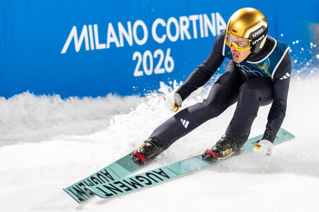 German ski jumper in gold helmet skiing on snow with Milano Cortina 2026 sign in background