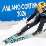 German ski jumper in gold helmet skiing on snow with Milano Cortina 2026 sign in background