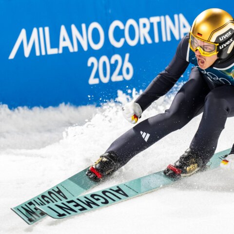 German ski jumper in gold helmet skiing on snow with Milano Cortina 2026 sign in background