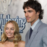 Margot Robbie and Jacob Elordi posing at the Wuthering Heights event, Robbie in a white corset dress and Elordi in a gray suit.