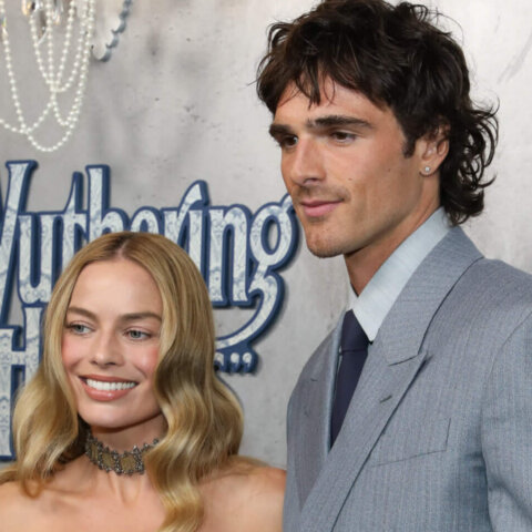 Margot Robbie and Jacob Elordi posing at the Wuthering Heights event, Robbie in a white corset dress and Elordi in a gray suit.