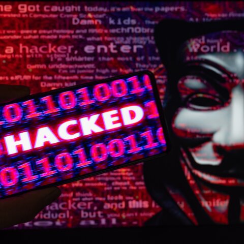 Hand holding smartphone displaying "HACKED" with binary code overlay and Guy Fawkes mask in background.