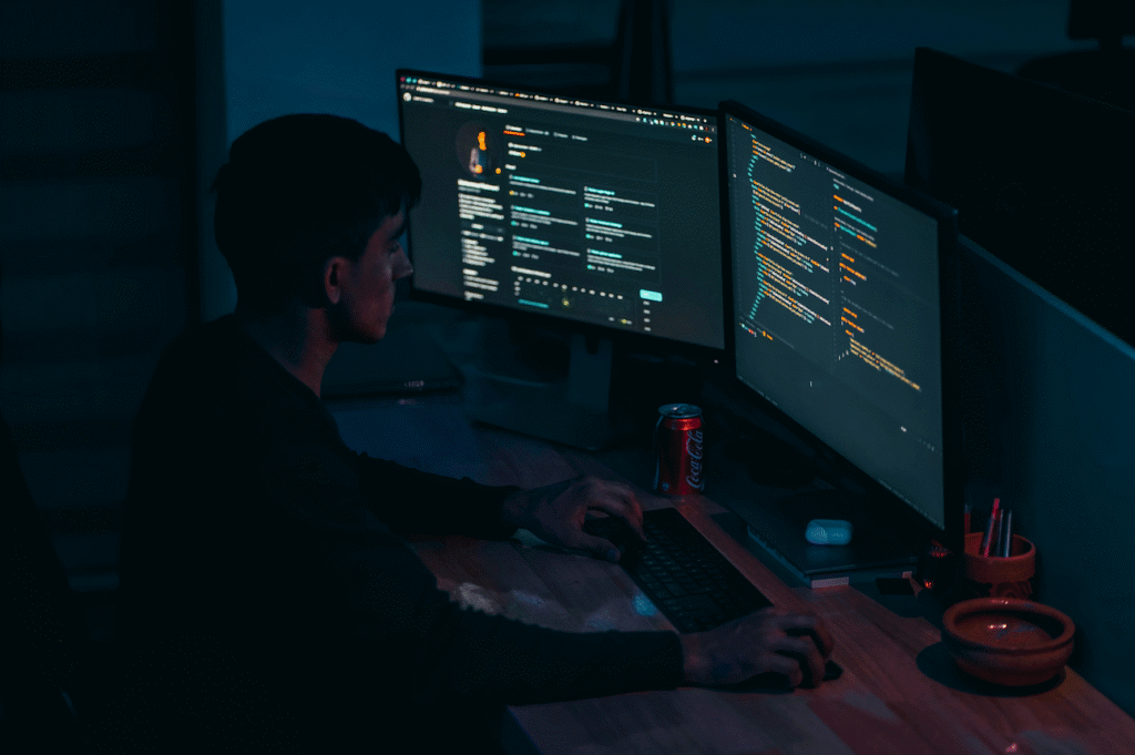 Person working on dual monitors displaying code and a profile page, with a Coca-Cola can on the desk.