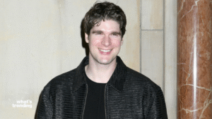 Man smiling wearing a black textured jacket and black shirt against a beige wall with a marble column