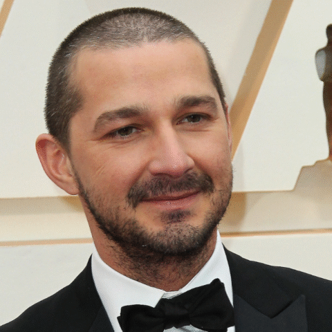 Shia LaBeouf in a black tuxedo with bow tie at an awards event with an Oscar statue in the background