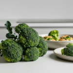 Fresh broccoli florets on a white kitchen countertop with plates of broccoli and food in the background