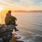 Man in green jacket sitting on cliff edge overlooking ocean at sunset with mountains in the distance