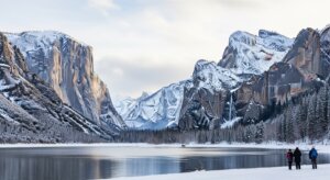 Three people walking near a snowy lake with snow-covered mountains and cliffs in the background at Yosemite National Park.