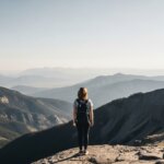 Person with backpack standing on mountain cliff overlooking layered mountain ranges under clear sky