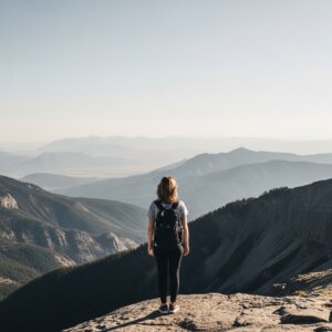 Person with backpack standing on mountain cliff overlooking layered mountain ranges under clear sky
