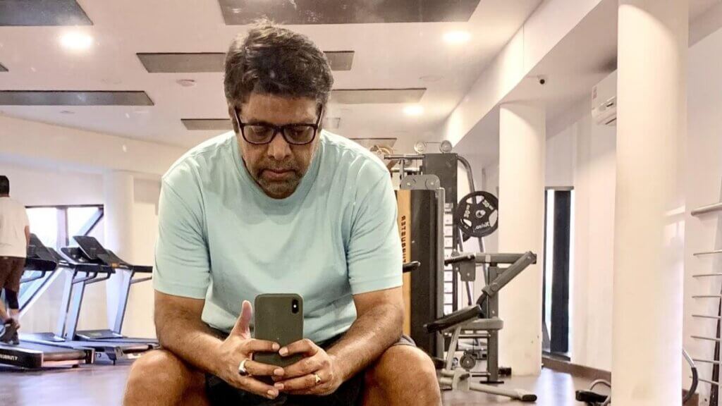 Man in glasses and light blue shirt sitting in gym taking a selfie with smartphone