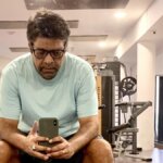 Man in glasses and light blue shirt sitting in gym taking a selfie with smartphone