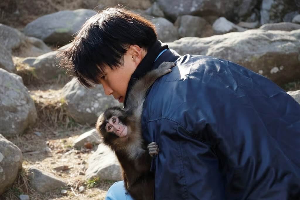 Young man in blue jacket hugging a small monkey outdoors among rocks