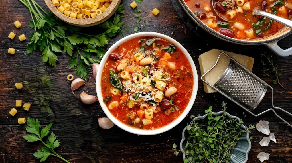 Bowl of minestrone soup with pasta, beans, and grated cheese surrounded by herbs, garlic, and a cheese grater on a wooden table