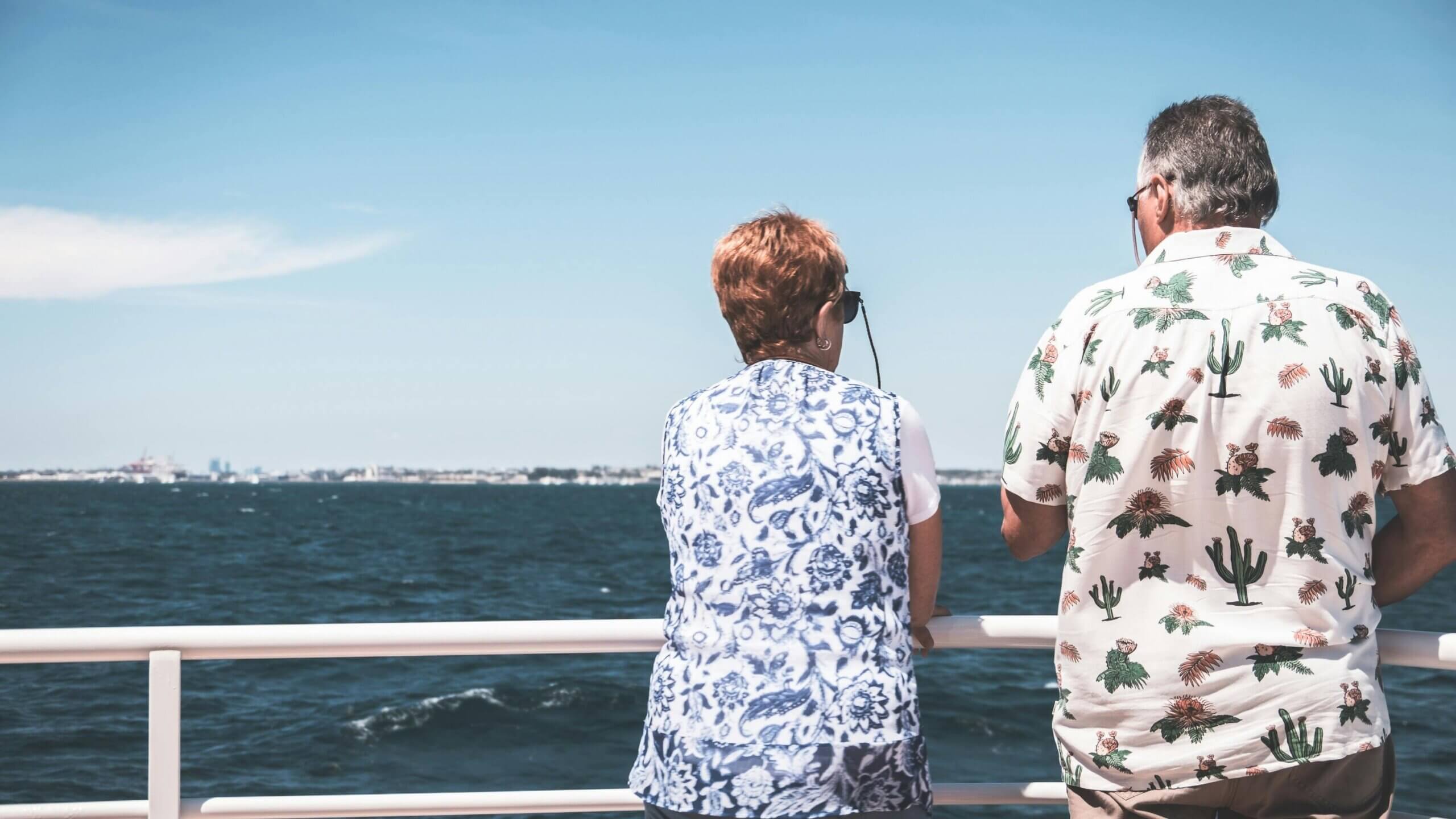 Two people wearing patterned shirts stand on a boat railing looking out over the ocean on a clear day.