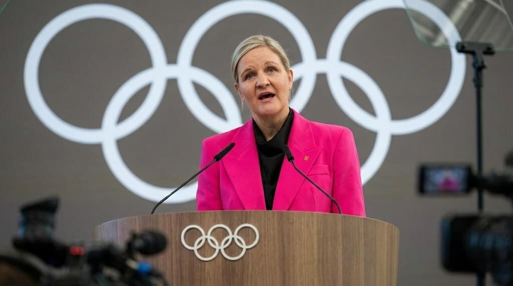 Woman in a pink blazer speaking at an Olympic podium with Olympic rings in the background