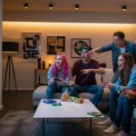 Group of six diverse young people playing video games together in a living room with snacks on the table.
