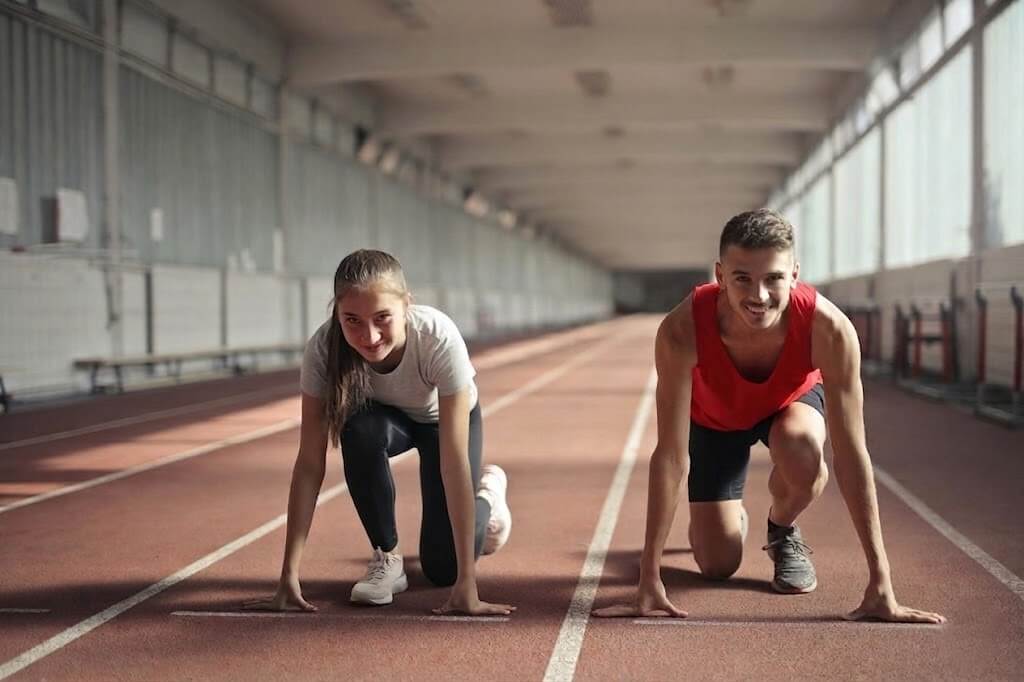 Two young athletes in starting positions on an indoor track, a woman in white and a man in red tank top.