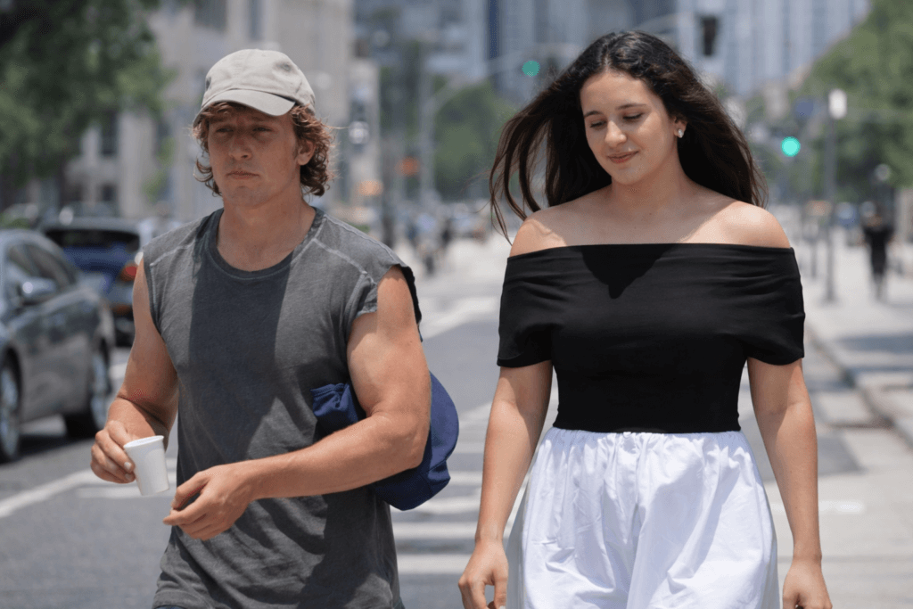 Young man in a gray sleeveless shirt and cap holding a cup walking beside a woman in a black off-shoulder top and white skirt on a city street.