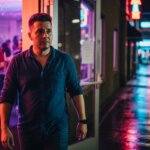 Man in blue shirt walking out of a neon-lit venue onto a wet city sidewalk at night