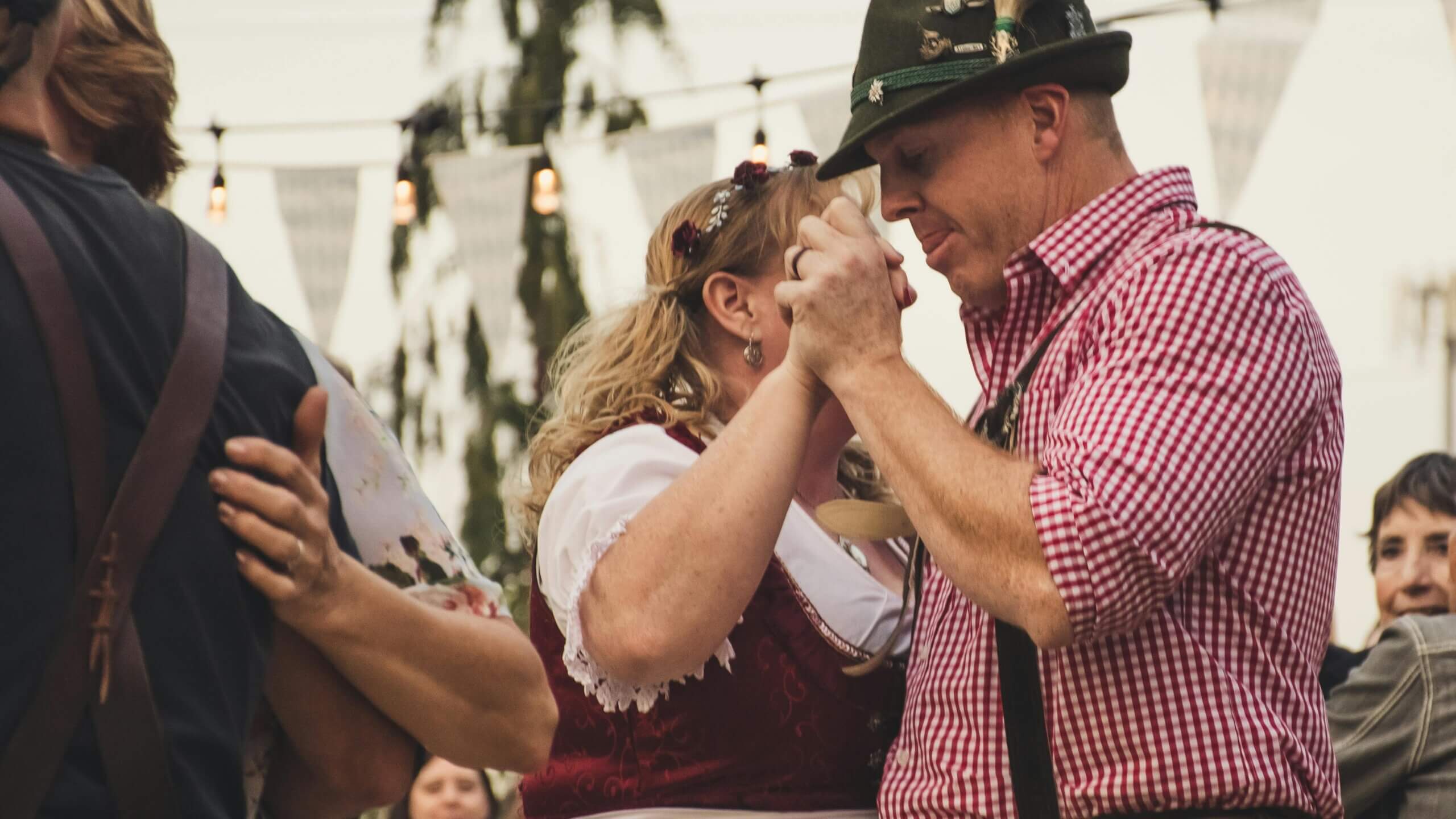 Couple dancing in traditional Bavarian clothing at an outdoor festival with string lights and bunting.