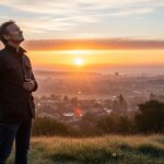 Man in black jacket standing on grassy hill at sunrise overlooking cityscape.