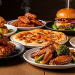 Steaming plates of fried chicken, chicken wings, pizza, and two burgers with sauce on a wooden table