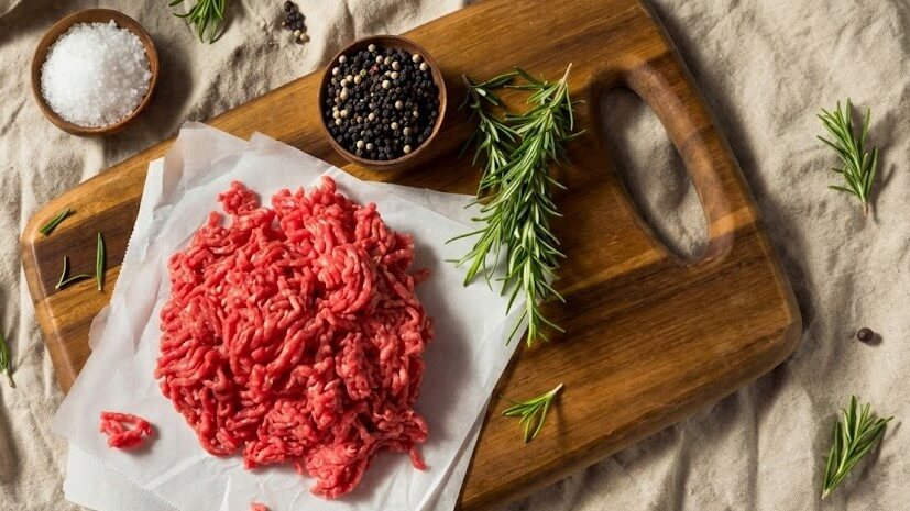 Raw ground beef on parchment paper with rosemary sprigs, salt, and peppercorns on a wooden cutting board