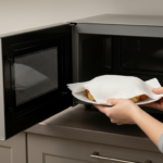 Hands placing a plate of food covered with a paper towel into a silver microwave oven in a kitchen.