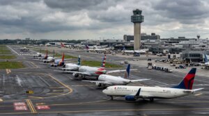 Delta, United, American, and Southwest airplanes lined up on a wet runway at an airport with a control tower under cloudy skies.